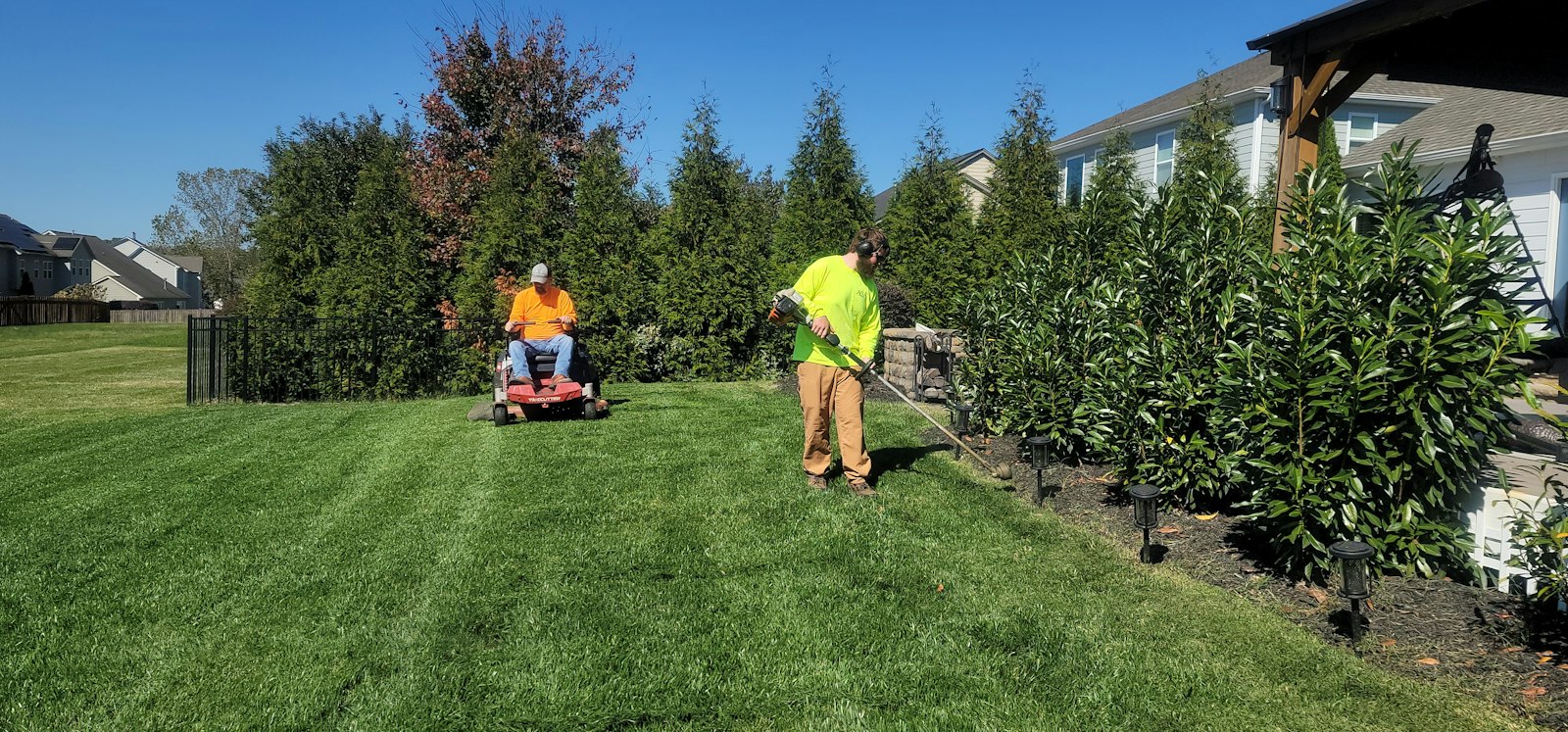 Two workers mowing a wide lawn with ride-on and push mowers